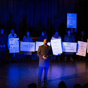 Actors on stage recreating the March on Washington and Dr. Martin Luther King, Jr.'s "I have a dream speech."  Actors hold signs that say "freedom" "we shall overcome" "fight for our rights" and more.