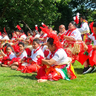 Tongan particants dressed in colorful attire seated on the grass and chairs.