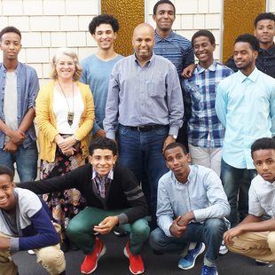 Group photo of East African All Stars. 11 young men and 3 adults standing in front of brick wall. 