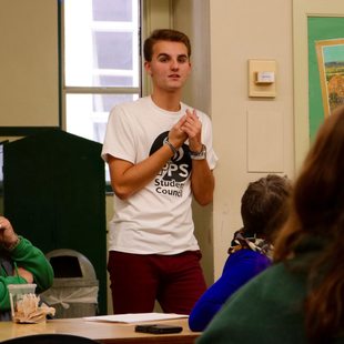 a high school student speaks to peers at a meeting while standing