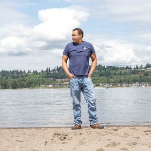 A black man stands on the bank of the Columbia River on a sunny day with blue skies.