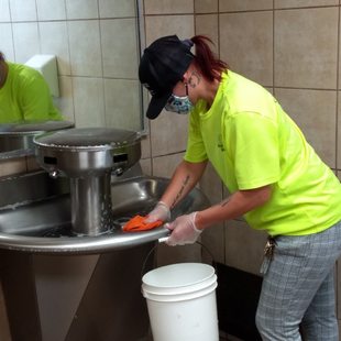 a sanitation worker cleans a sink in a public restroom while wearing their mask