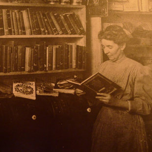 A sepia-tone photograph of a woman reading a big, leather-bound book in front of a shelf of other big, leather-bound books.