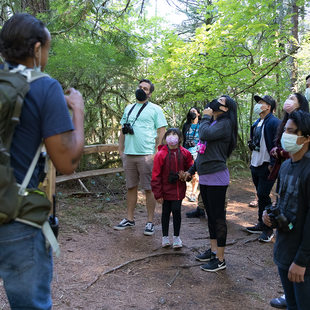 A group of hikers wearing masks listen to a nature educator wearing a large backpack.