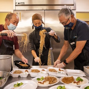 Three cooks place long strings of noodles in to-go food containers