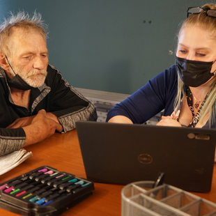 Older white man on the left and blonde woman with mask on the right, looking at a laptop on a table