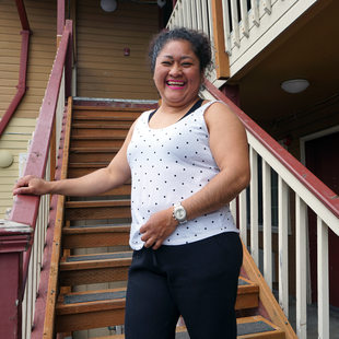 Latina woman in white polka dot tank top and black pants smiling and standing at foot of exterior stairs