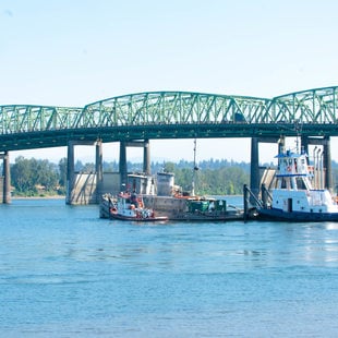 Two tugboats maneuver two derelict boats in the Columbia River, with the Interstate Bridge in the background.