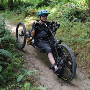 Max Woodbury rides an adaptive mountain bike down a dirt path in woods