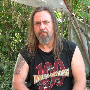 Man with long hair wearing a Harley-Davidson shirt with a tarp and bike behind him