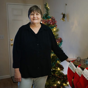 Woman with short hair in black shirt in front of Christmas tree. Her hand is resting on a table with Christmas stockings hanging off the edge.