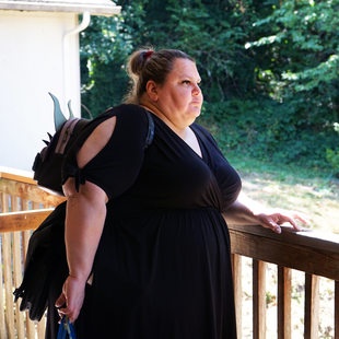 Woman standing at a wooden railing looking into the distance, wearing a black dress.
