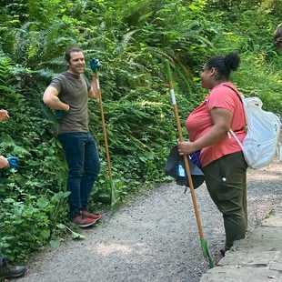 Four young people talk on a path in a forest, there are lush ferns lining one side of the path and a concrete wall on the other