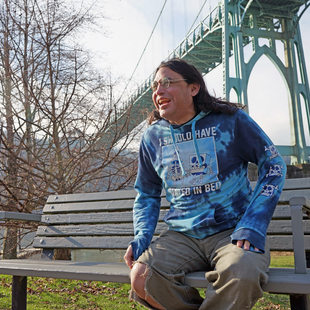 Man in blue printed sweatshirt sitting on a bench, laughing, with the St. Johns bridge in the background.