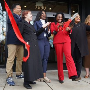 Eight people just after cutting a large red ribbon in front of an apartment building. A woman in the center in a red suit holds oversized red scissors