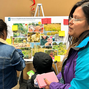 Two women and a child look at a board with different pictures of outdoor spaces. Sticky notes are attached to the board with comments from community members.