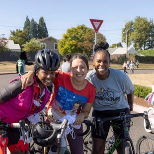 Three adults pause to take a group photo. The adult to the left is wearing a bike helmet and cycling gear.