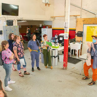 A group of people in a semi-circle watching a person speak, the background is an industrial warehouse