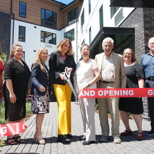 Eight people posing for a photograph just after cutting a red "grand opening" ribbon with giant scissors.