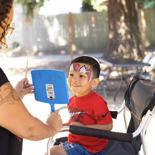 A young light brown skinned boy with face paint smiles while looking in a mirror the face painter holds up for him.