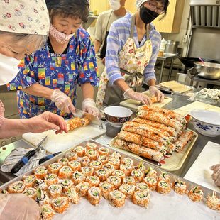 Three Japanese women wearing masks and colorful patterns prepare sushi in a small kitchen.