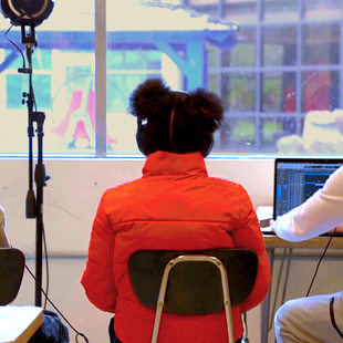 A group of young people sitting in a classroom with headphones on in front of a laptop. A microphone on a stand is nearby.