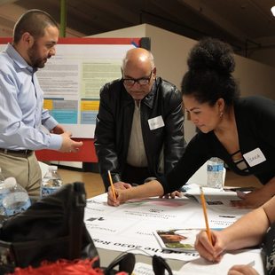 A diverse group of people stand around a table working on posters during a community meeting. They wear name tags and a large bulletin board stands in the background. 