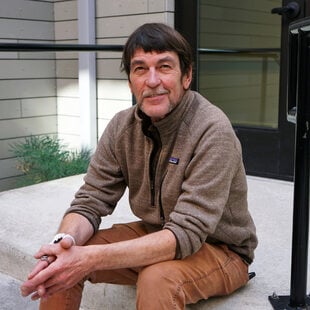 Middle aged man siting on the steps in an apartment building courtyard. He's wearing a brown sweatshirt and brown pants.