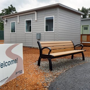 Bench in front of a grey bungalow, with a Welcome sign next to it. 