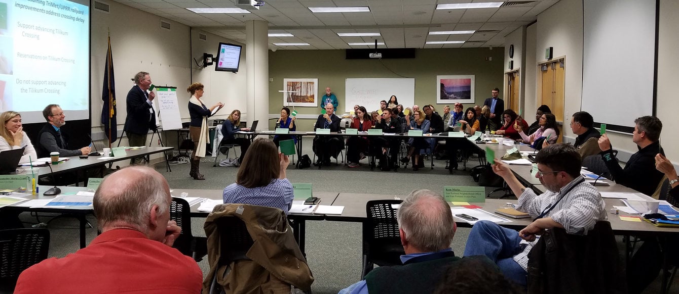 Two people presenting to a room full of people sitting around tables with a display screen in the background.