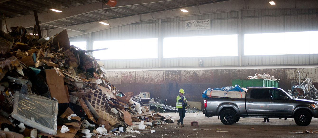 transfer station staff person in a yellow vest stands near big pile of garbage