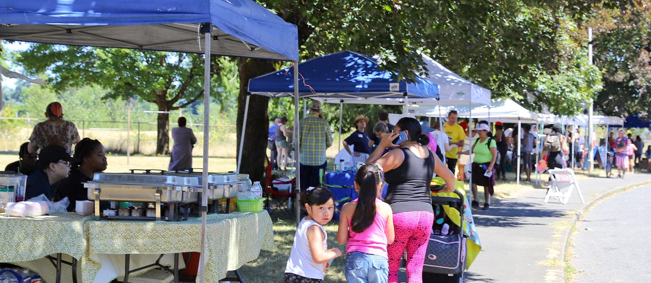 Families walk past park vendors on a tree-lined street.