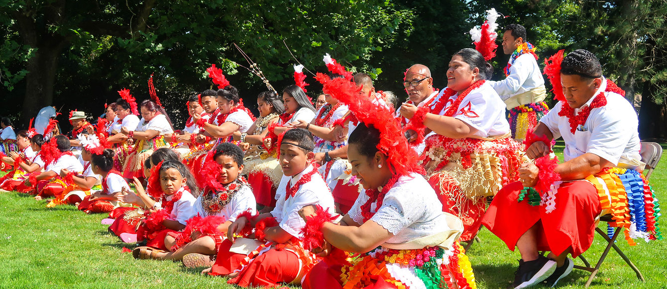 Tongan particants dressed in colorful attire seated on the grass and chairs.