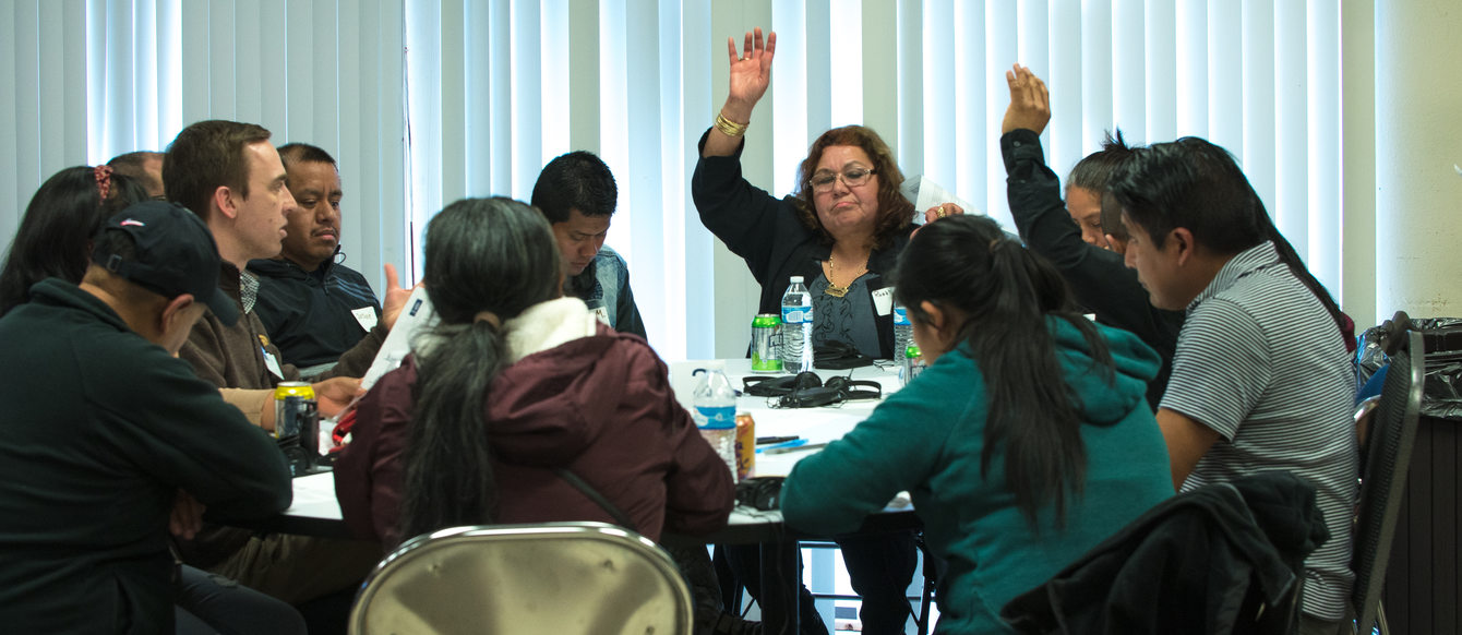 People sitting around a table, some taking notes, some raising their hands to speak