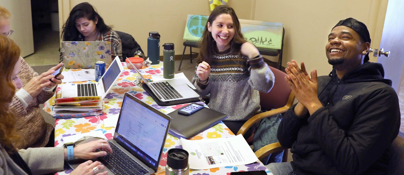 Five people huddle over laptops at a table in a tiny office space. Two of them are laughing. The only man in the room wears a broad smile and throws his head back.