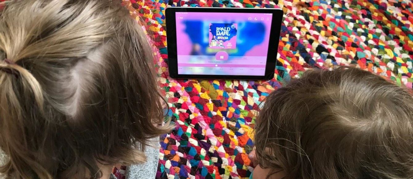 two little kids listen to an audio book on a tablet while laying down on a rug on the floor