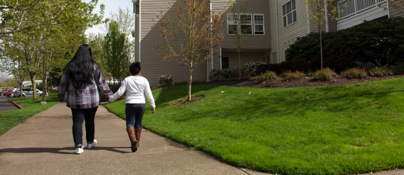 an adult and a child walk hand-in-hand on a sidewalk in front of an apartment complex