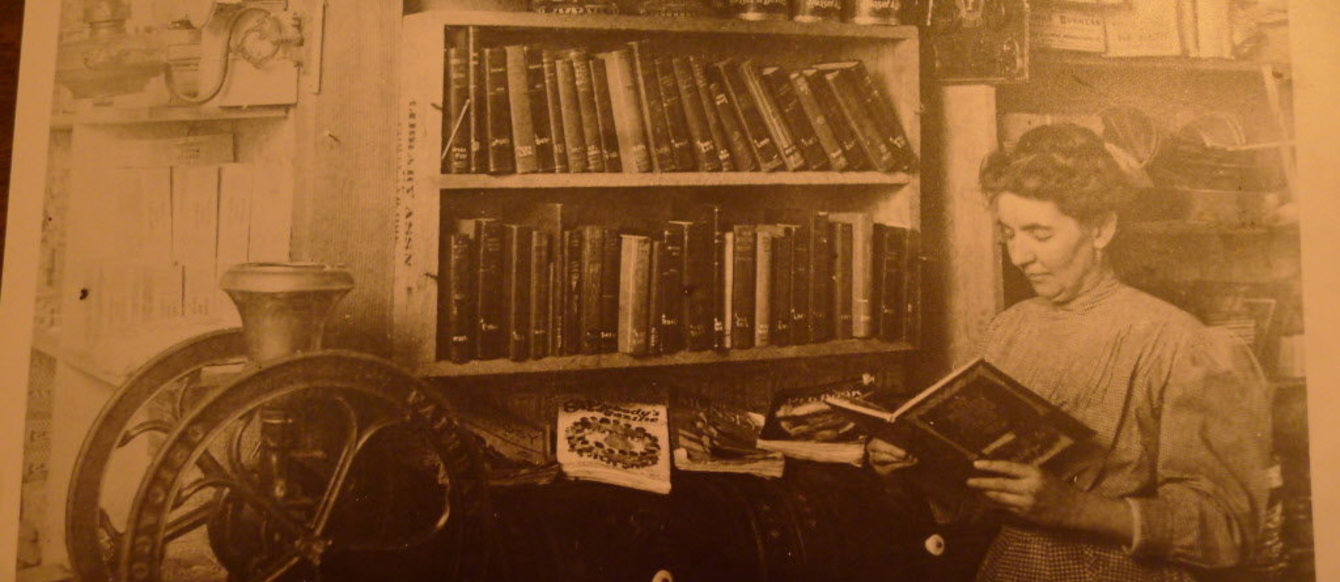 A sepia-tone photograph of a woman reading a big, leather-bound book in front of a shelf of other big, leather-bound books.