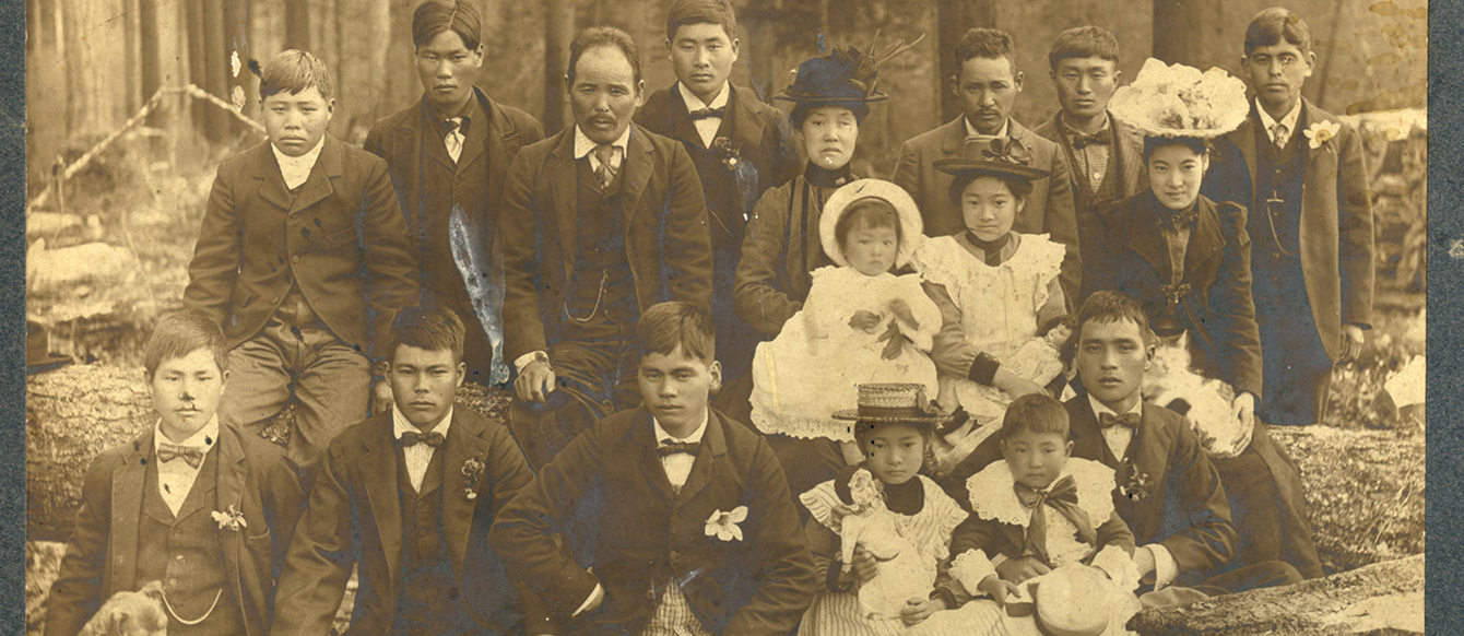 A sepia-toned photo of group of about 20 Japanese people in suits and dresses posing for a photo in a forest.