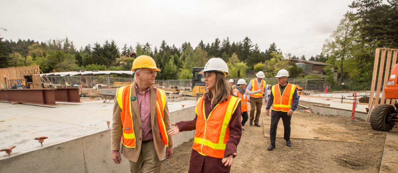 Don Moore and Heidi Rahn, wearing hard hats and safety vests, walk through the elephant prairie construction site at the Oregon Zoo. More people wearing hard hats and safety vests are seen in the background.