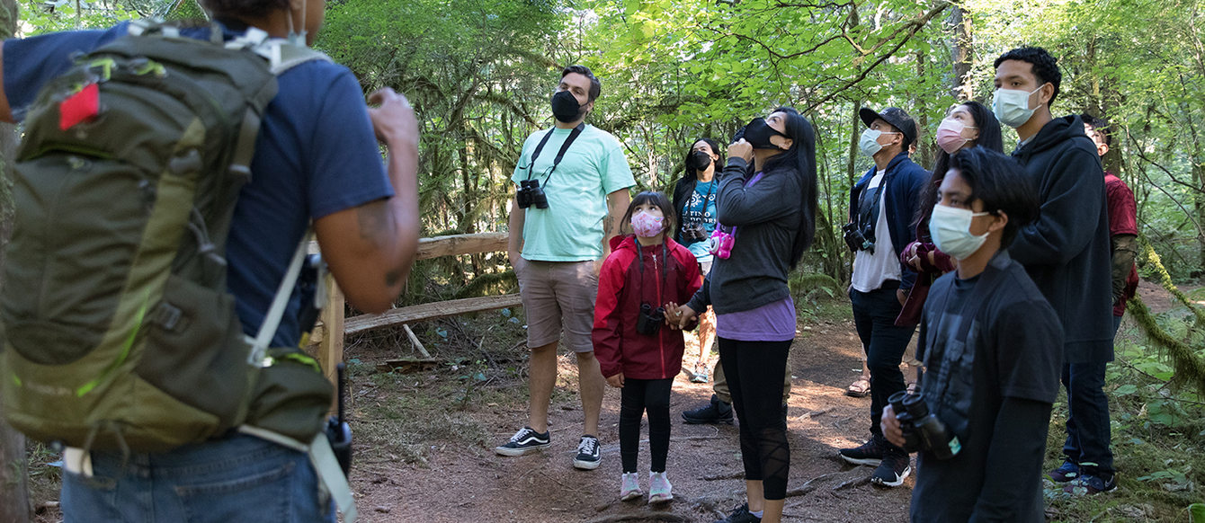 A group of hikers wearing masks listen to a nature educator wearing a large backpack.