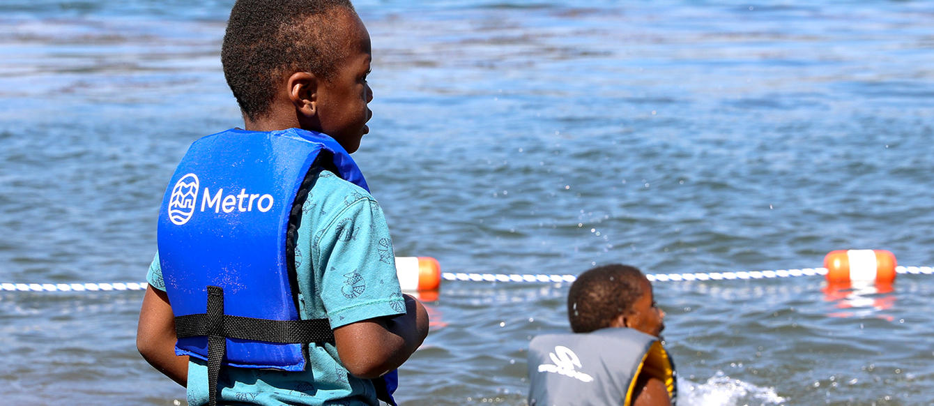 A young Black boy stands wearing a life jacket with Metro printed on the back while another young Black boy wearing a life jacket splashes in the Columbia River.