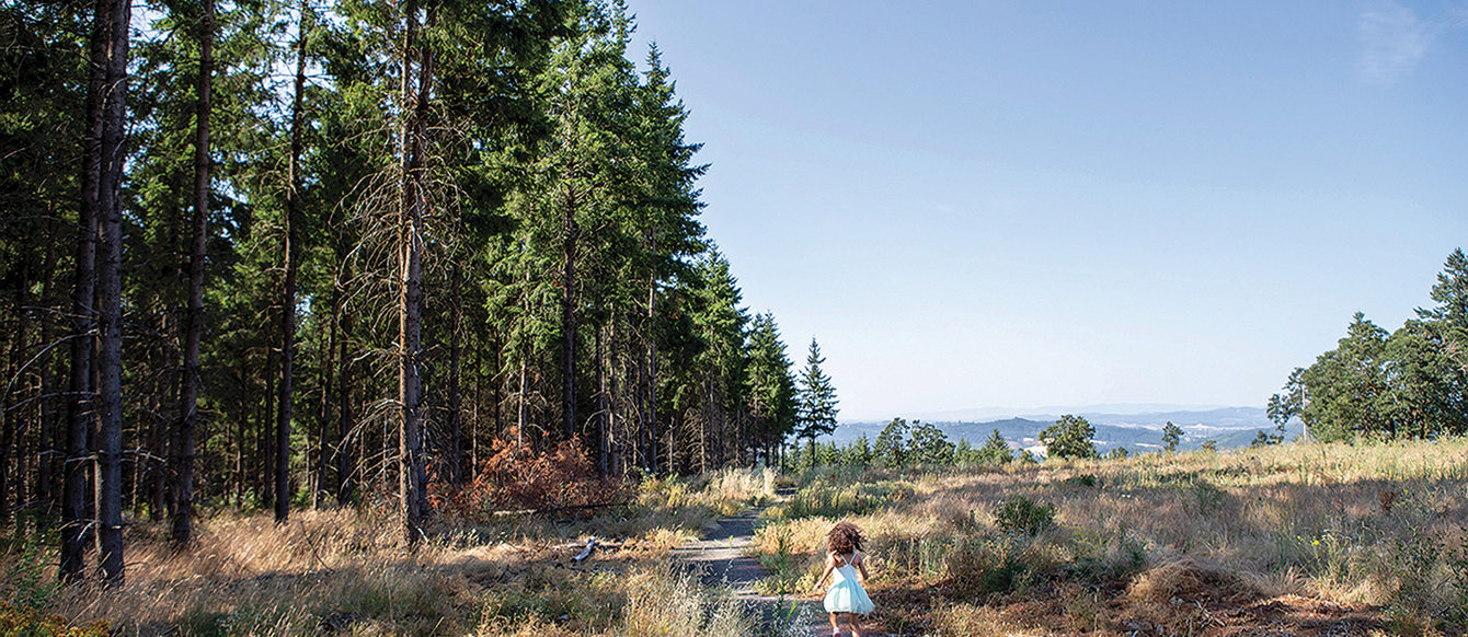A young child wearing a blue dress runs along a trail with tall trees on one side and an open prairie on the other.