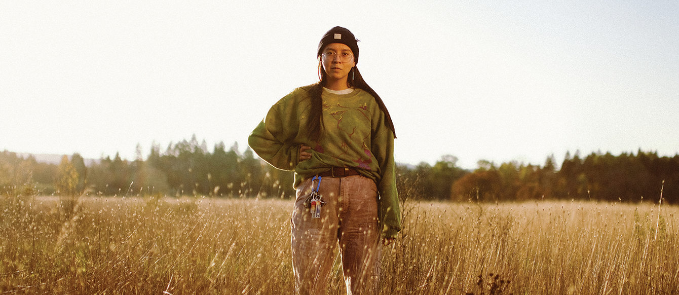 A 20-something young person with long black hair stands in a field of dried grass that comes up to their waist.