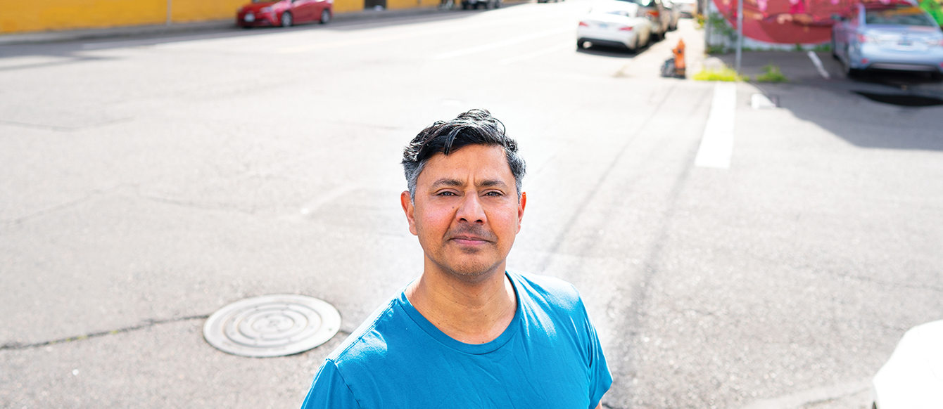 A man stands looking at the camera. Behind him is a wide street with several large warehouses. There is no greenery.