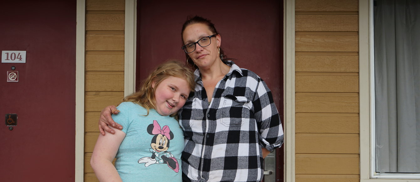 A woman wearing glasses and a black-and-white plaid shirt stands with a child wearing a Minnie Mouse t-shirt and pink striped pants in front of the entrance door to an affordable housing unit