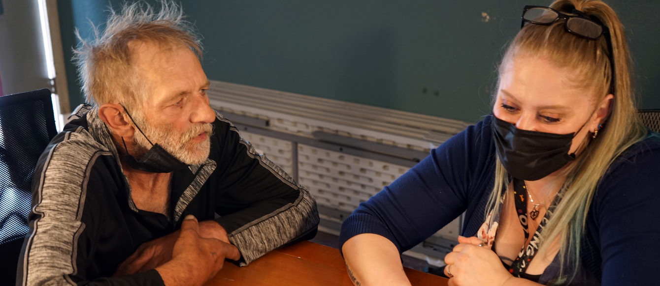 Older white man on left with younger blonde white women on the right. Sitting at a table looking at a laptop.