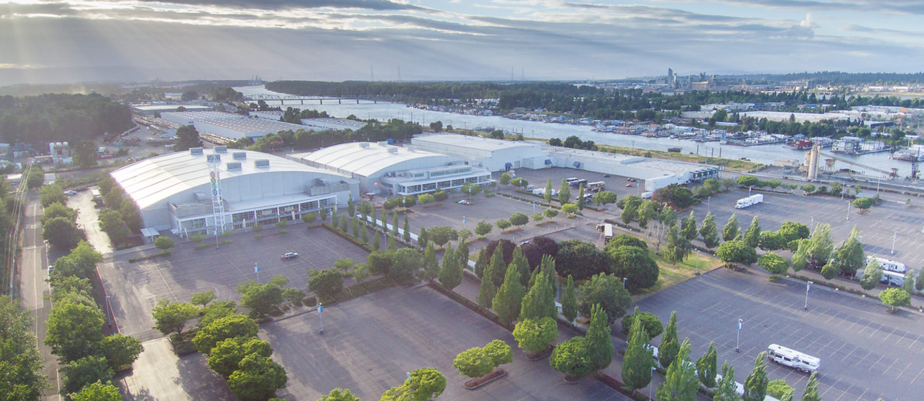 Aerial view of Portland Expo Center, showing the full property, buildings, and parking lots. The view is looking west toward the setting sun, with rays of sunlight streaming through the clouds onto the Expo Center property