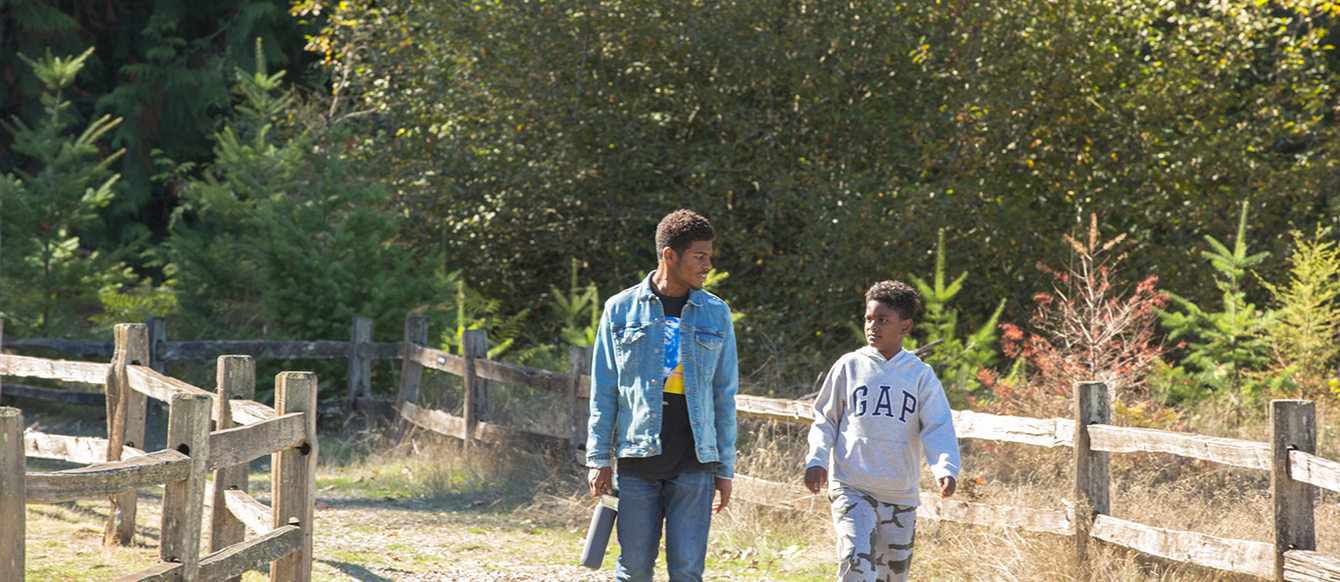 A young Black man and a Black child talk as they walk along a fenced trail in a park.