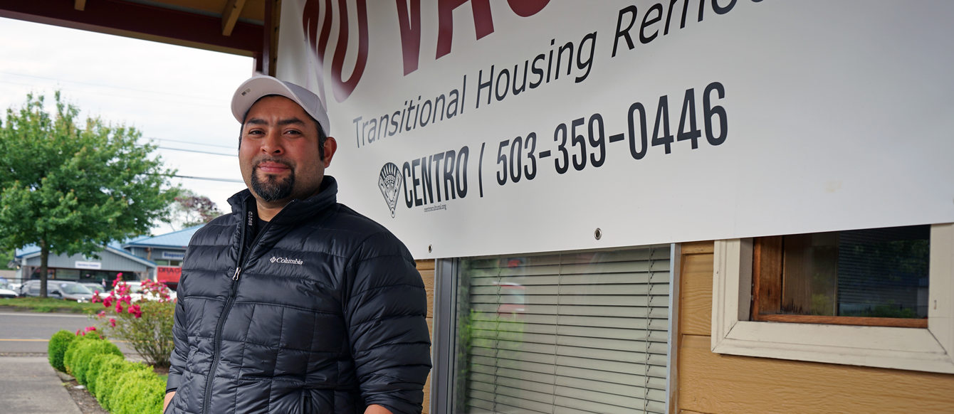 Latino man standing in front of a building with a sign that says "transitional housing" and has a phone number